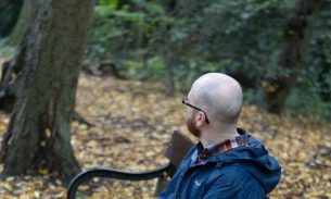 A man sits on a woodland bench with his back turned to the camera